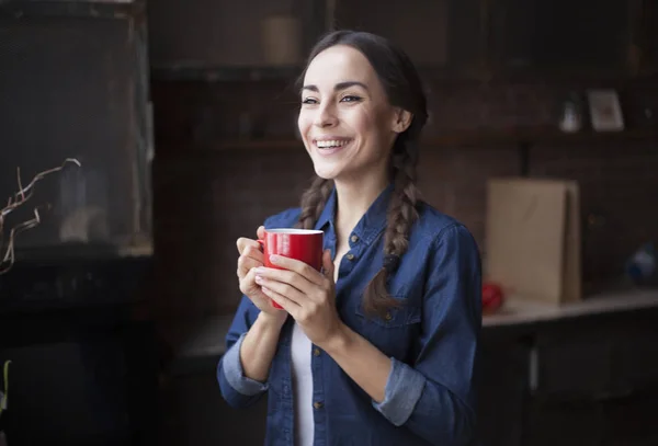 Портрет очень красивой юной брюнетки в джинсовой рубашке с красной чашкой кофе в руках. Девушка улыбается на кухне дома . — стоковое фото