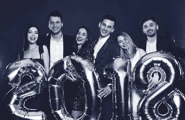 New Year in the circle of best friends. Group of cheerful young people in beautiful evening dresses and costumes carrying gold colored numbers and smiling in camera. Black and white