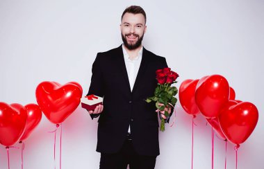 Zarif siyah takım elbise yakışıklı gülen adam elinde bir hediye kutusu ve kırmızı güller ile kameraya izole beyaz arka plan üzerinde görünüyor..