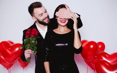 Akşam güzel bir sevgi dolu gülümseyen çift elbise, bir erkek bir kadın için bir hediye yapar ve gözleri kırmızı balonlar bir kalp şeklinde arka planı kapatır.