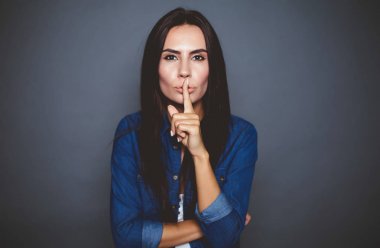 Bu bizim sırrımız olacak. Güzel bir konsantre kadın kot gömlek onun parmağı dudakları için izole bir gri arka plan üzerine koyar.