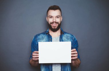 Her şey tam size göre. Yakışıklı genç gülümseyen adam kot gömlek fotoğraf makinesi gri bir arka plan üzerinde beyaz bir sayfa gösterir. Reklam alanı.