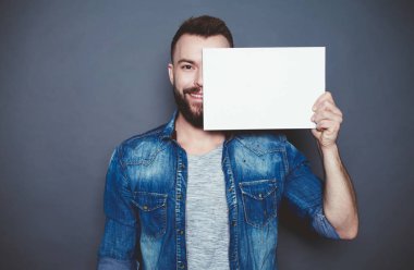 Her şey tam size göre. Yakışıklı genç gülümseyen adam kot gömlek fotoğraf makinesi gri bir arka plan üzerinde beyaz bir sayfa gösterir. Reklam alanı.