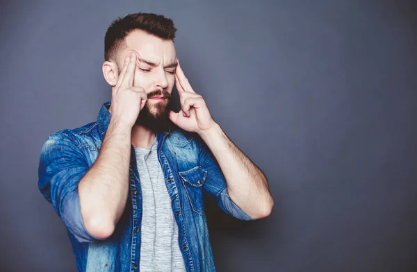 After stress. A young bearded man in a denim shirt with a headache on a gray background.