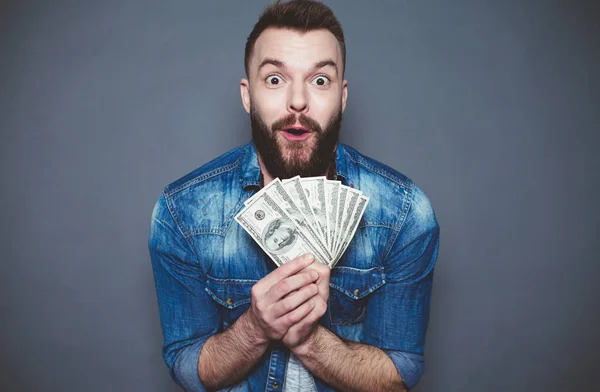 Monetary winnings. Young joyful man in a denim shirt holds a lot of dollars in the hands and shocked looking at the camera on a gray background.