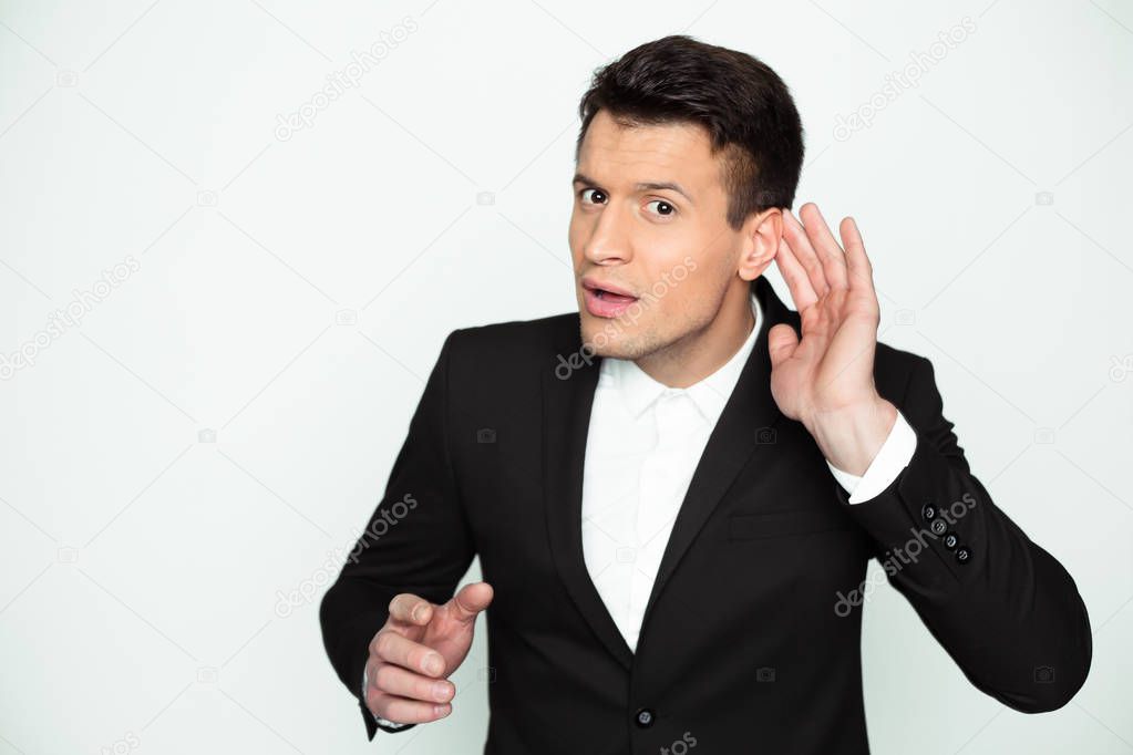 What are you saying? A young man in a black jacket holds his hand near the ear with a questioning emotion on his face.