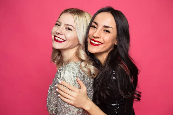 Friends forever. Close up photo of Two happy young beautiful smiling girlfriends in little black dresses posing and having fun on a pink background.