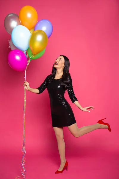 Girl with helium balloons. Beautiful smiling stylish woman in a little black dress with colorful helium balloons on a pink background.