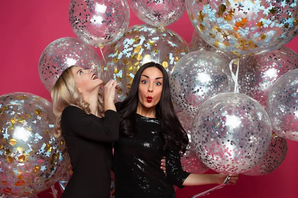 Holiday with helium balloons. Two beautiful smiling modern women in little black dresses are posing in front of the camera on a pink background with large helium balloons.