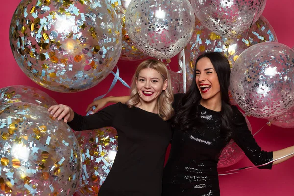 Holiday with helium balloons. Two beautiful smiling modern women in little black dresses are posing in front of the camera on a pink background with large helium balloons.