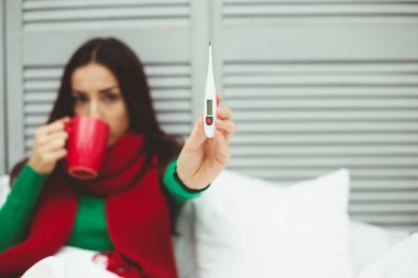 Çok yüksek vücut sıcaklığı. Kırmızı fularlı genç ve hasta bir kadınla bir termometre ile sıcaklık ölçer. Sağlık ve hastalık kavramı.