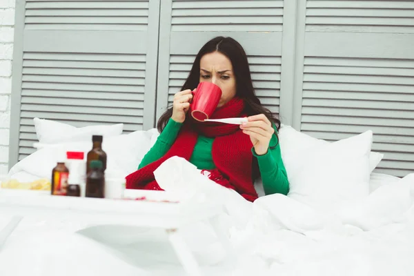 Very high body temperature. A young sick woman in a red scarf measures the temperature with a thermometer. The concept of health and disease.