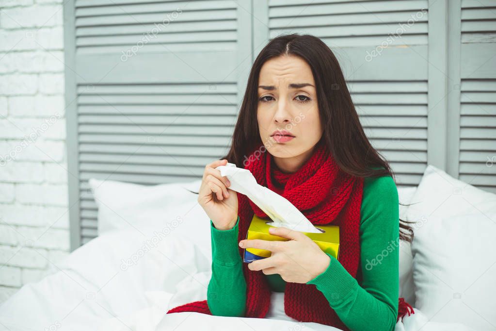 Close up portrait of a young sick woman with a runny nose in a red scarf lying on the bed at home and treated. The concept of health and disease.