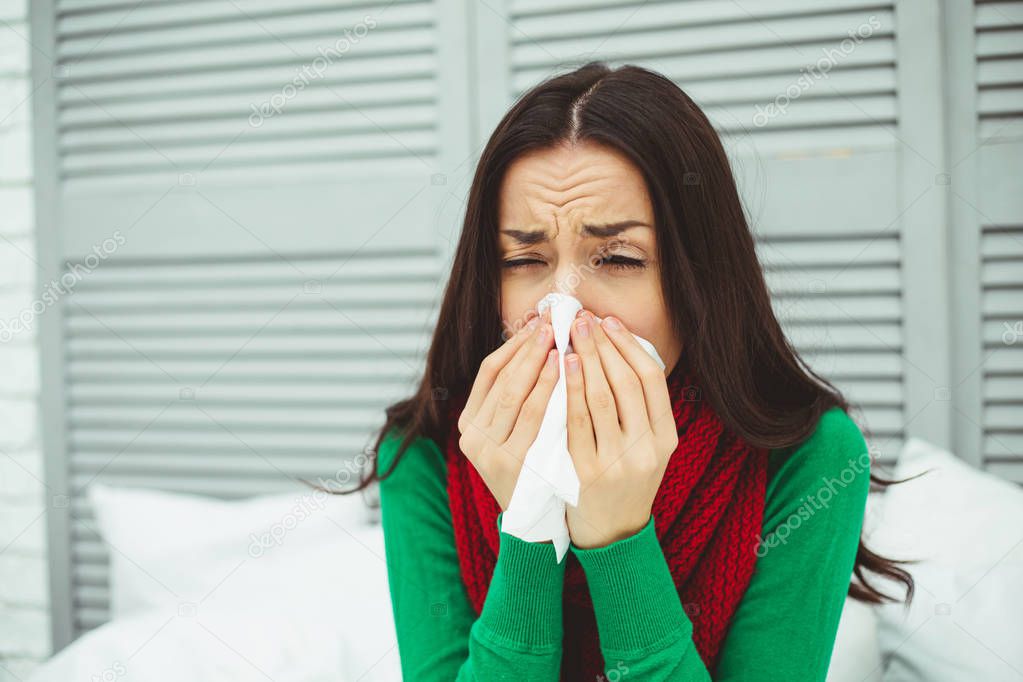 Close up portrait of a young sick woman with a runny nose in a red scarf lying on the bed at home and treated. The concept of health and disease.