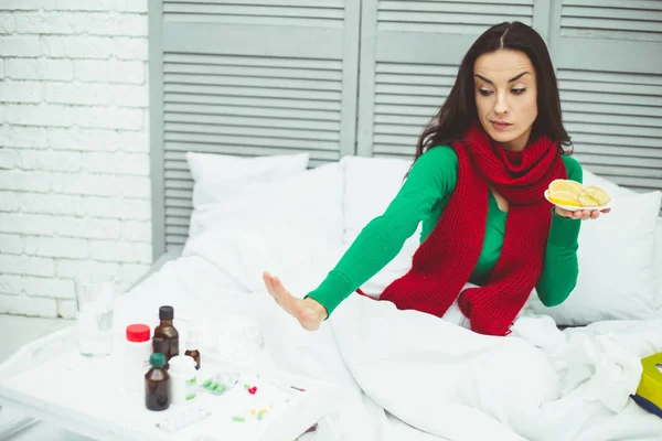 It is better to be treated without pills. A young smiling woman in a red scarf at home on the bed shows that tea and fruits with vitamins are better for treatment than tablets.