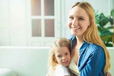 Güzel gülümseyen sarışın anne ve kızı kucaklamak ve kanepede evde ve bak kameraya eğlenin. Anne ve kızı kavramı.