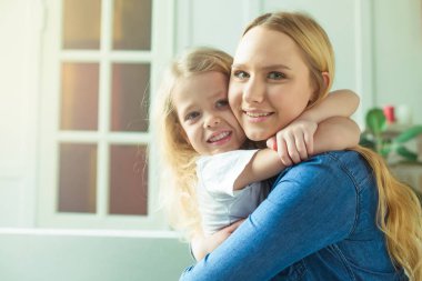 Güzel gülümseyen sarışın anne ve kızı kucaklamak ve kanepede evde ve bak kameraya eğlenin. Anne ve kızı kavramı.