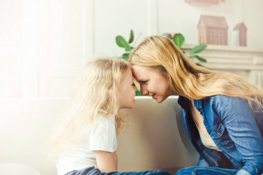 Gülümseyen güzel sarışın anne ve kızı alınlarına birbirlerine evde kanepede oturan dokun. Anneler günü. Kadınlar Günü. 8 Mart.