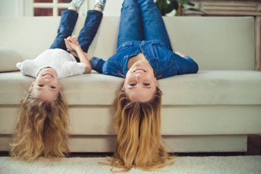 Anne, sorun değil! Güzel gülen sarışın anne ve kızı kanepede başlarını aşağı asmak ve kameraya bak.