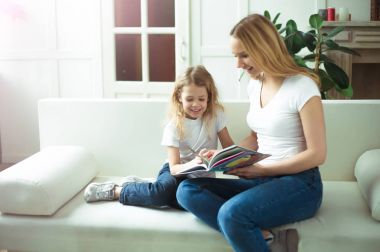 Güzel gülümseyen sarışın anne ve kızı bir kucaklama içinde kanepede oturuyorsun. Anne bir hikaye kızı ya da bir peri masalı bir kitap okuyor.