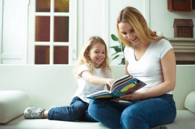 Güzel gülümseyen sarışın anne ve kızı bir kucaklama içinde kanepede oturuyorsun. Anne bir hikaye kızı ya da bir peri masalı bir kitap okuyor.