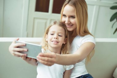 Anne ve kızı. Güzel gülümseyen sarışın anne ve kızı evde kanepede kucaklamak ve telefonda kendilerini fotoğraf çekmek veya selfies yapmak.