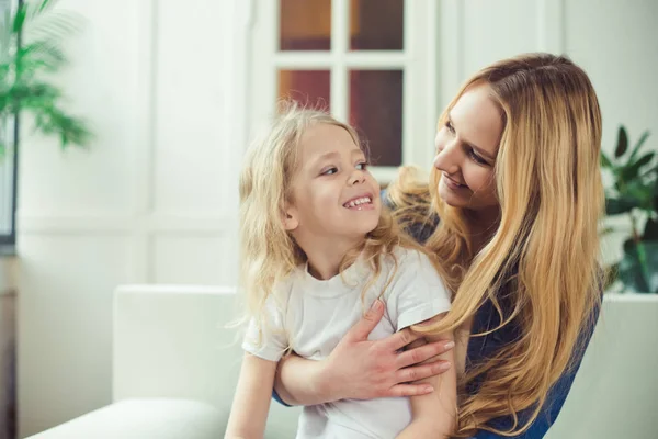 Smiling and happy mom and daughter are hugging each other at home on the couch. Mom and daughter  have fun.