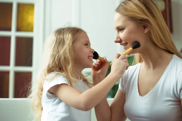 Mom teaches her daughter how to use makeup. Beautiful smiling blond mother and daughter are sitting on the sofa at home. Little girl holding a brush for powder.