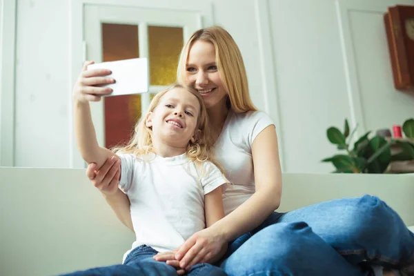 Mom and daughter. Beautiful smiling blond mother and daughter embrace on the sofa at home and take pictures of themselves on the phone or make selfies.