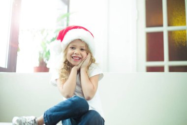 Gülümseyen güzel sarışın küçük kız evde kanepede oturan ve kameraya bakarak Noel Baba şapkası. Yeni yıl ve Noel.