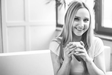 Evde bir kanepede oturan ve sıcak kahve içme güzel genç sarışın bir kadın portresi.