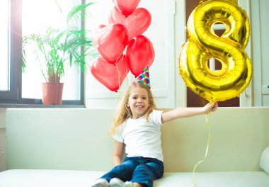 Mutlu güzel sarışın gülümseyen şapka şenlikli, küçük kız ve helyum balon sekiz rakamı şeklindeki evde doğum günü kutluyor.