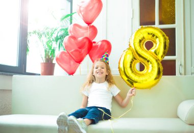 Mutlu güzel sarışın gülümseyen şapka şenlikli, küçük kız ve helyum balon sekiz rakamı şeklindeki evde doğum günü kutluyor.