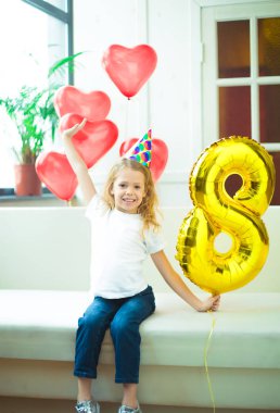 Mutlu güzel sarışın gülümseyen şapka şenlikli, küçük kız ve helyum balon sekiz rakamı şeklindeki evde doğum günü kutluyor.