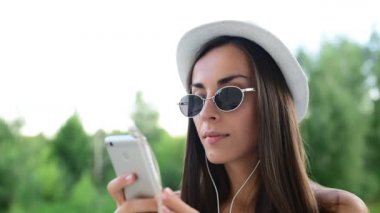Şapka ve güneş gözlüğü ile bir telefon ve onun favori müzik dinlerken kulaklık çok güzel neşeli kız arkadaş telefon ile iletişim.