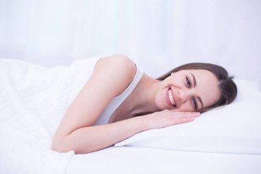 Genç güzel gülümseyen kadın evde beyaz yatakta yatarken uykudan uyanma.