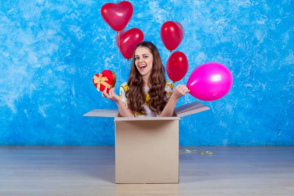 Happy Birthday. Funny smiling young girl sitting in a cardboard box with a gift and balloons and looking at the camera in casual clothes.
