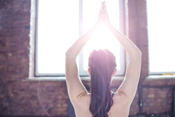 Rear view of a young woman practicing yoga in the gym on the background of a window in the sunlight. Healthy lifestyle. Yoga class