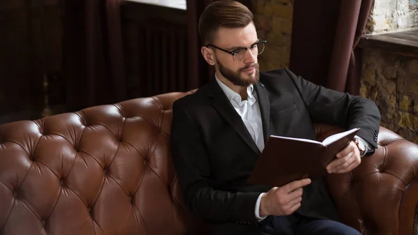 Confident handsome bearded businessman in a smart suit and glasses reading a book or check the reports on the work of sitting on the couch in the office.