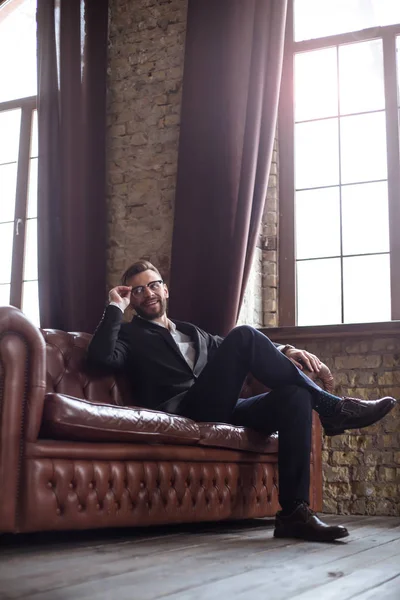 Handsome stylish confident bearded businessman in a smart suit and glasses sitting on a sofa in the office.