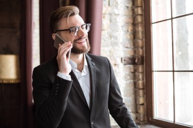 Takım elbise ve ofis pencere yakınındaki telefonda konuşan gözlük yakışıklı işadamı.