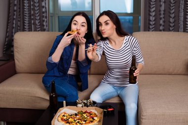 Kanepede arkadaşlarınızla sakin ol. İki güzel genç kız evde film izlemek ve rulo ve pizza yemek kanepede gülümseyen.