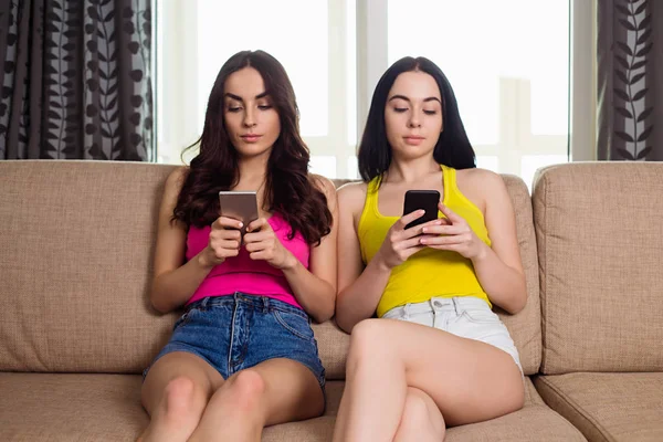 Duas Amigas Irmãs Bonitas Com Telefones Nas Mãos Sentadas Sofá — Fotografia de Stock