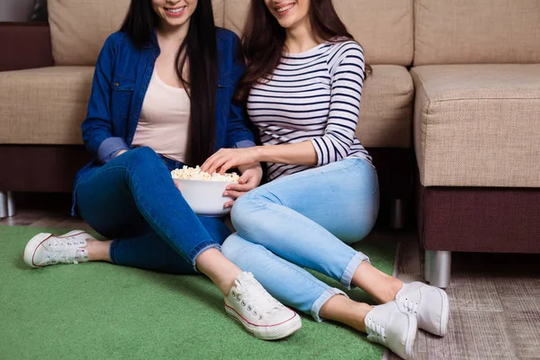 Two attractive girl friends at home watching TV or film and eat popcorn.