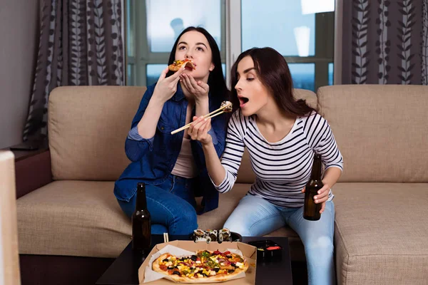 Relax on the couch with friends. Two beautiful young smiling girlfriends at home on the couch watching movies and eating rolls and pizza.