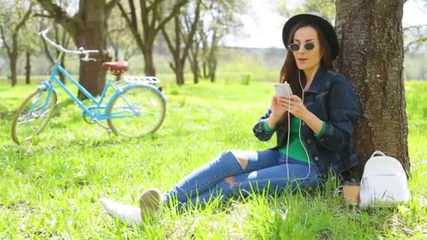 愉快和愉快的美丽的时髦女人在帽子和太阳镜听音乐与电话在城市公园和有乐趣在复古自行车背景 — 图库视频影像
