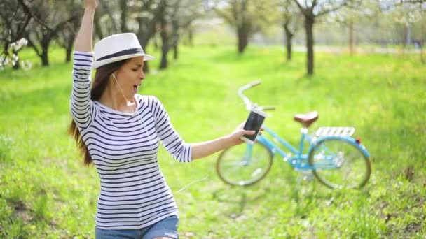 明るく 美しい若い女性の帽子とカジュアルを着る幸せな都市公園とレトロなバイクの背景に踊りで携帯電話で音楽を聴く — ストック動画