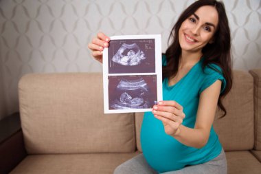 Bir ultrason resmini tutan ve kameraya gösteren kanepede oturan gülümseyen güzel hamile kadın. Çocuk bekliyor