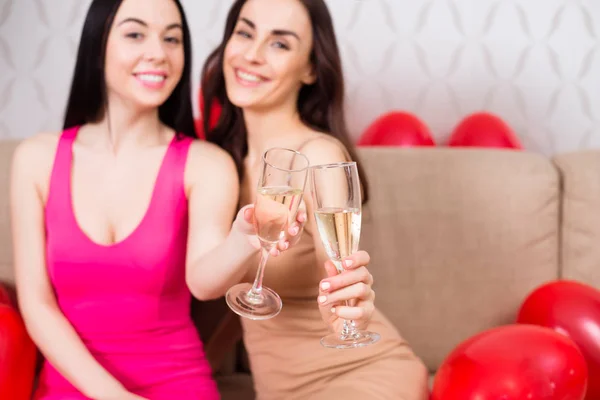 Hen-party. Two beautiful cheerful girls in bright evening dresses drink champagne and rest at a party sitting on the couch.