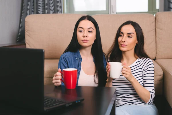 Two smiling beautiful women watching a movie or talking on skype in a laptop and drinking coffee or tea